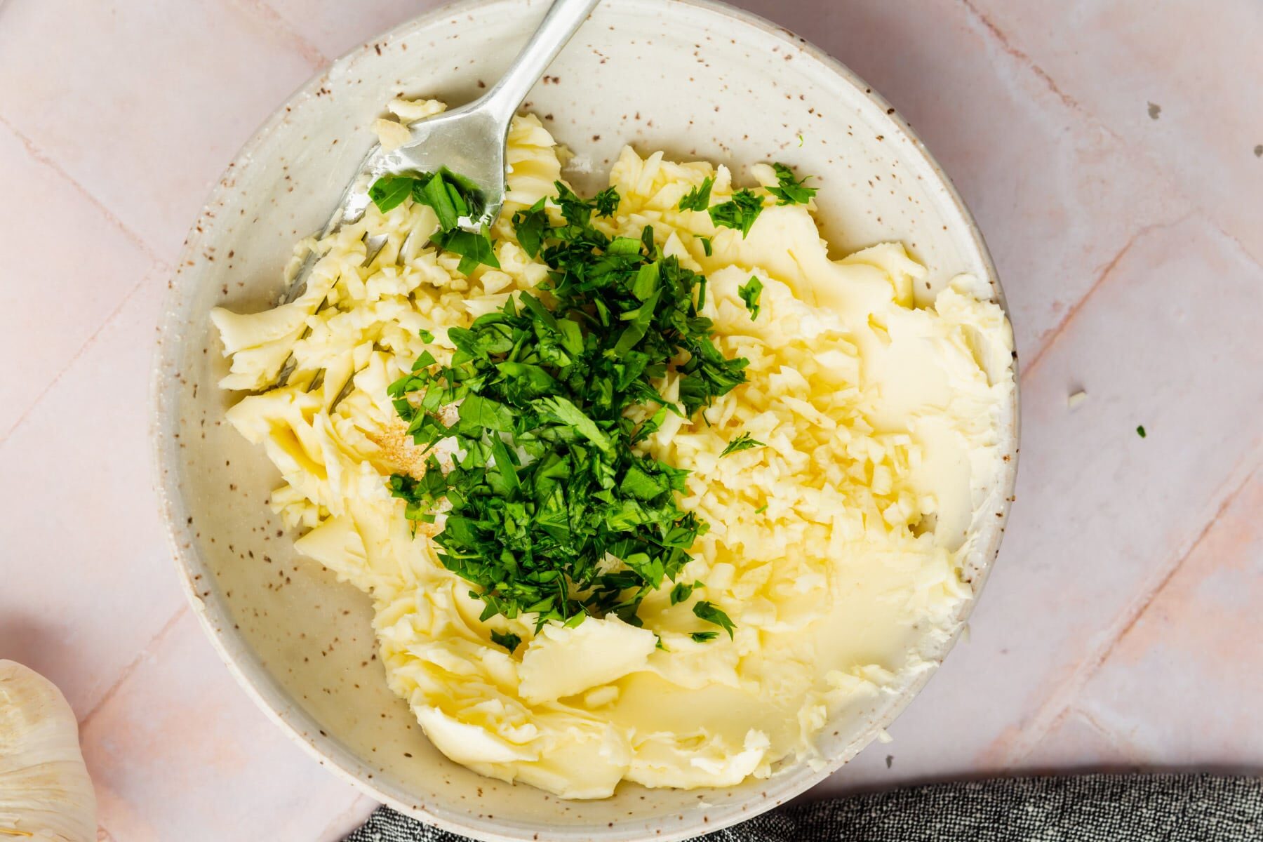 A bowl with butter, garlic powder, minced garlic and fresh parsley about to be mixed with a fork.