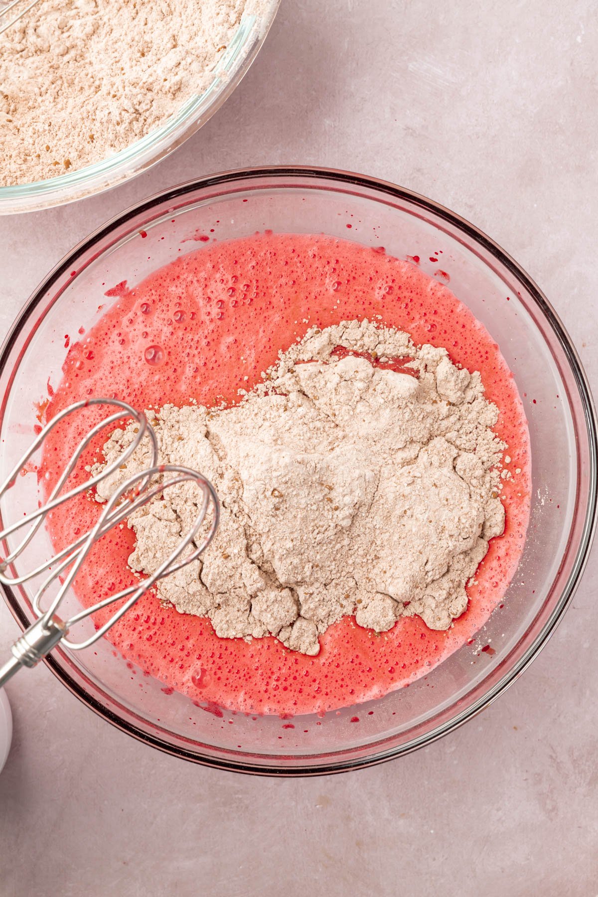 Red cake batter with brown dry ingredients in a glass mixing bowl with two whisk beaters.