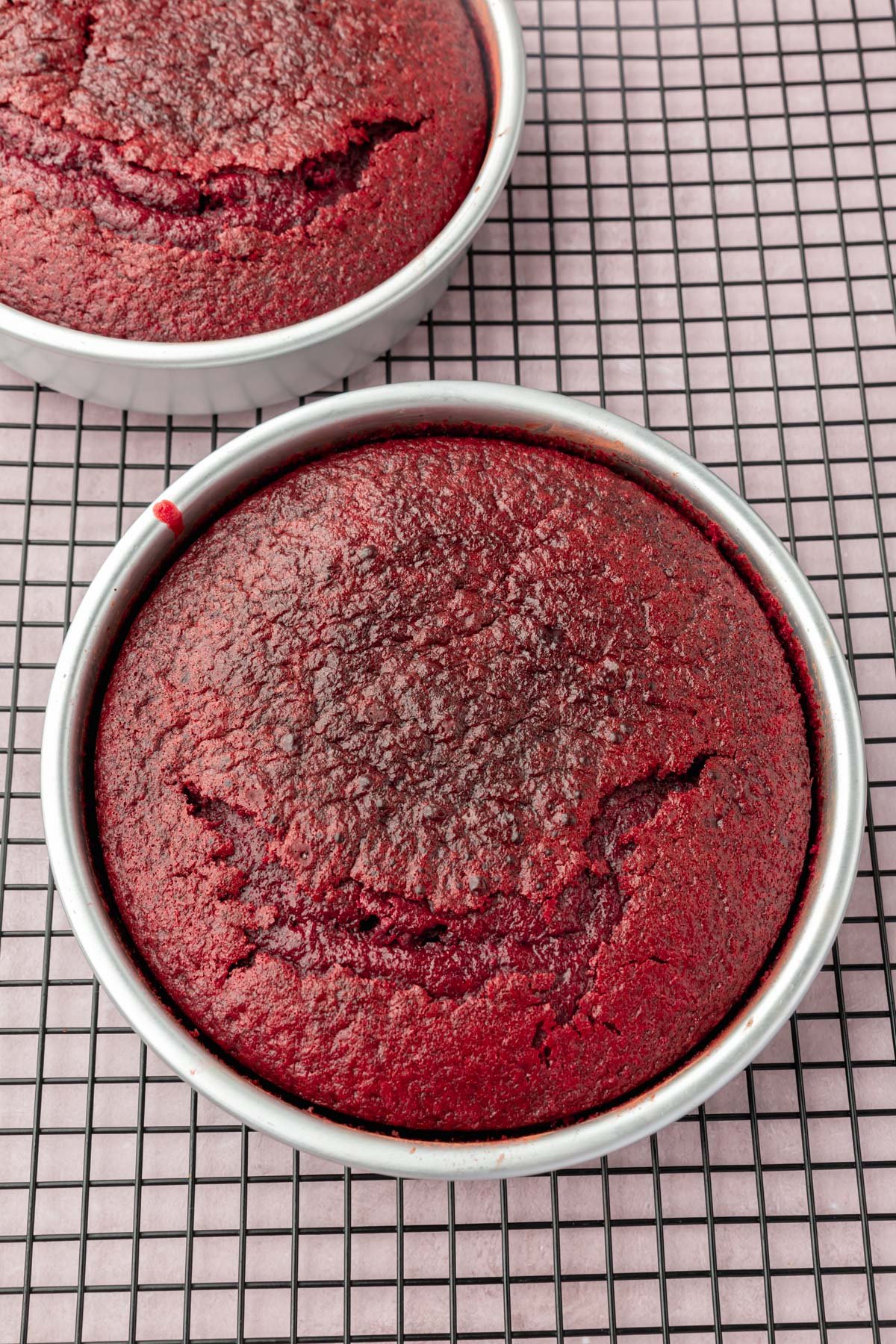 Two red velvet cakes in their pans on a wire cooling rack.