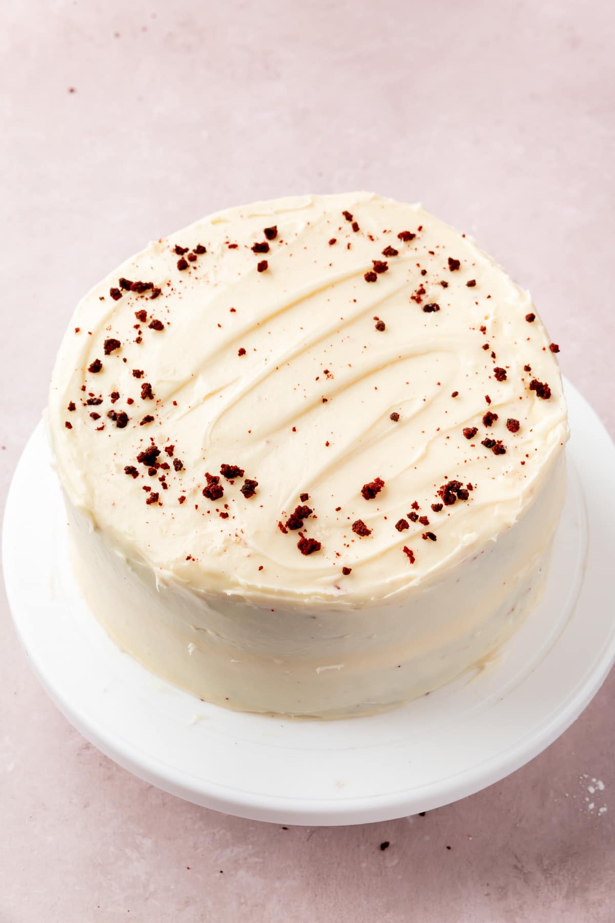 A red velvet cake frosted with cream cheese frosting and sprinkled with red velvet cake crumbs on top of a white cake stand.