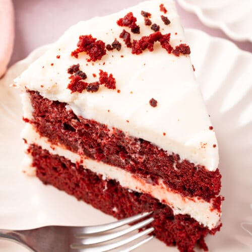 A slice of gluten-free red velvet cake on a plate frosted with cream cheese frosting and topped with a sprinkle of cake crumbs.