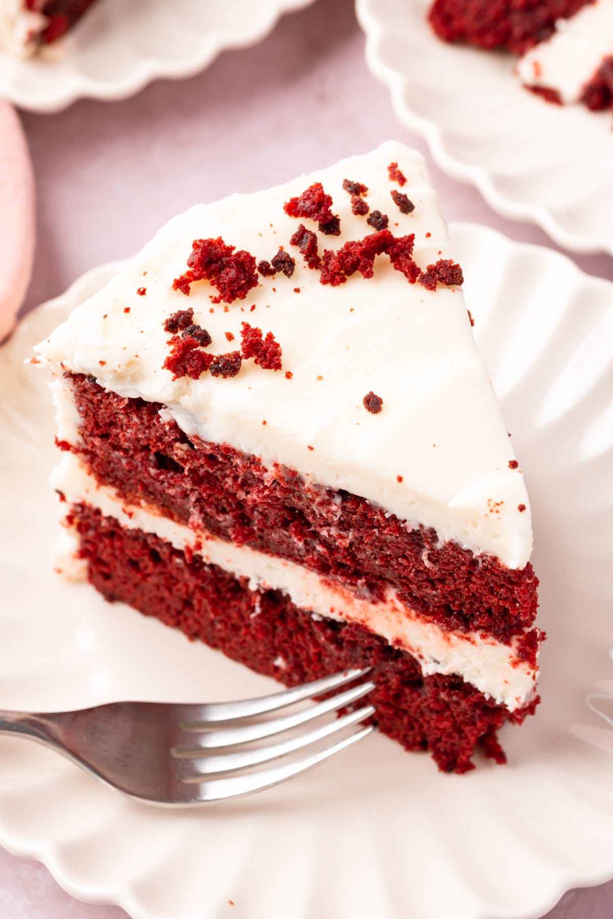 A slice of gluten-free red velvet cake on a plate frosted with cream cheese frosting and topped with a sprinkle of cake crumbs.