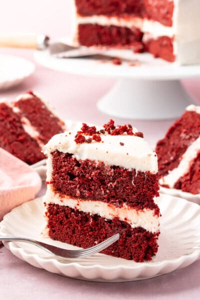 A slice of red velvet cake on a plate with a fork with more slices behind it.