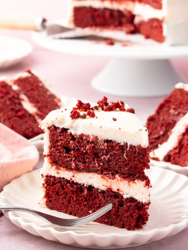 A slice of red velvet cake on a plate with a fork with more slices behind it.