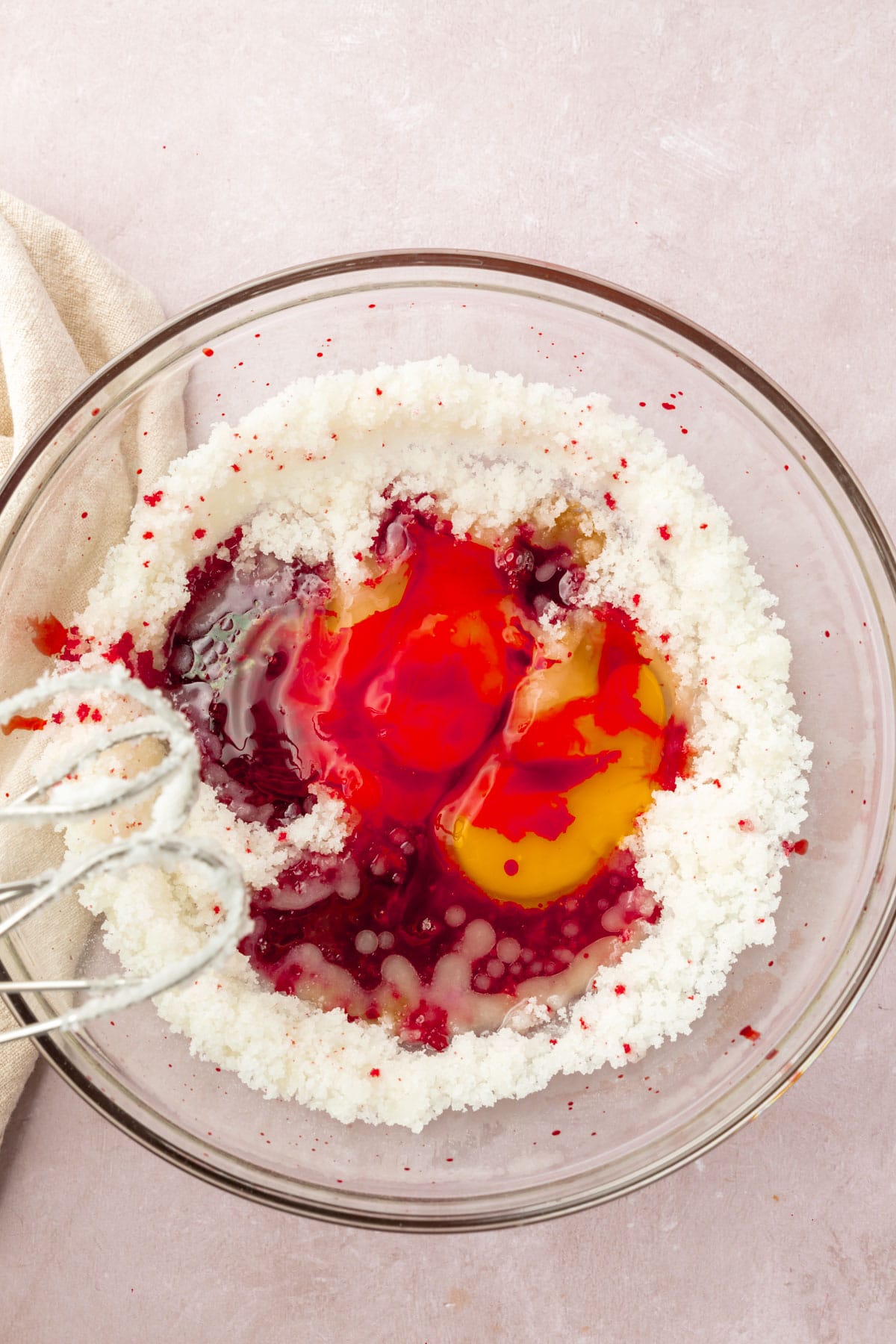 A bowl of granulated sugar, red food dye, vanilla, and eggs with two beaters.