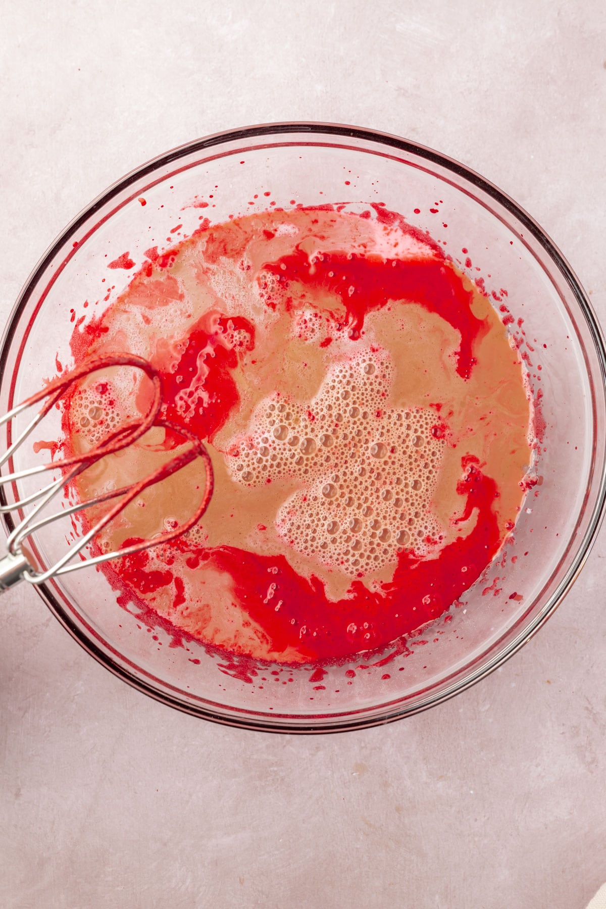 Red cake batter, coffee, and buttermilk in a glass mixing bowl with two beaters.