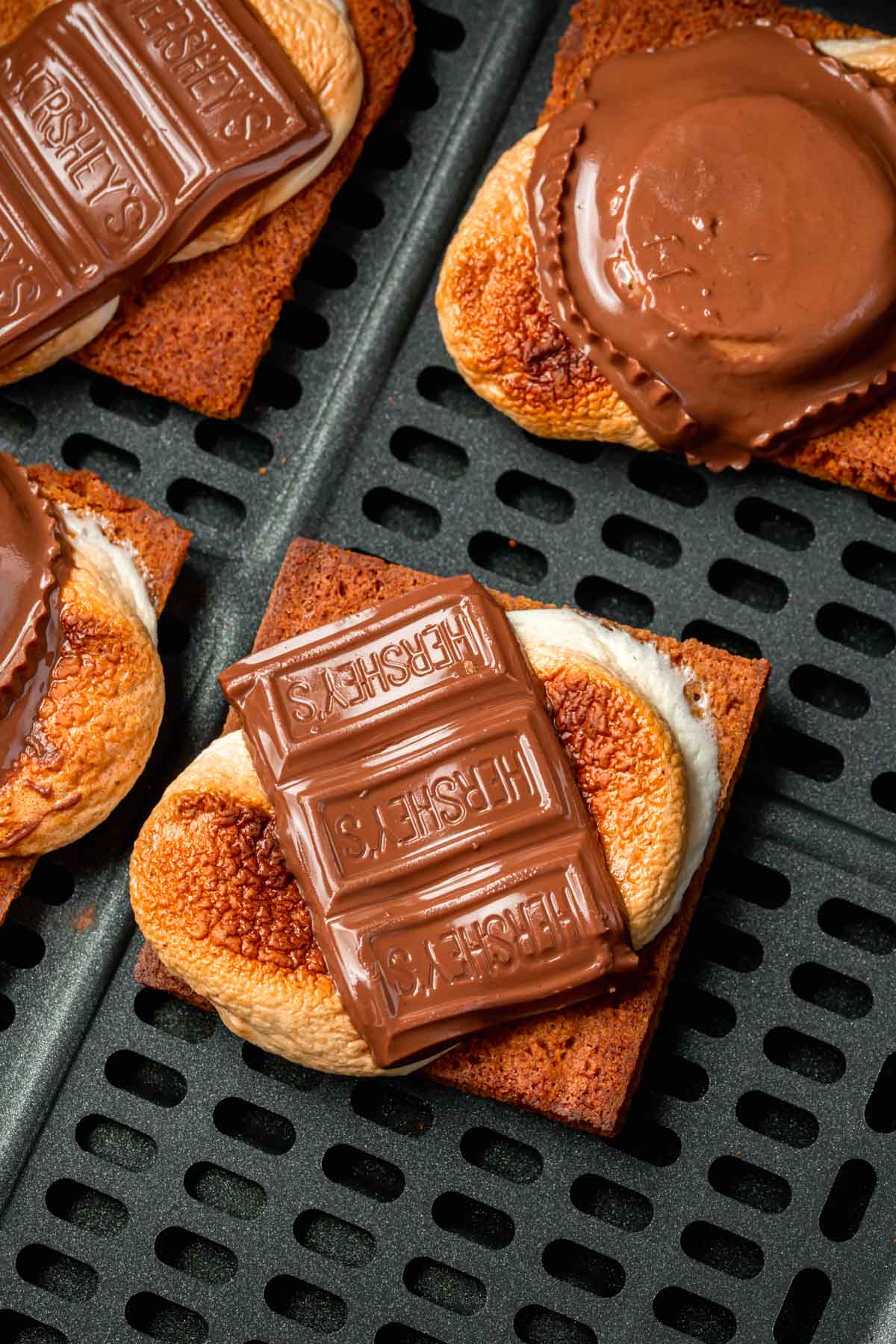 An air fryer basket with four open-faced s'mores in it featuring hershey bars and peanut butter cups.