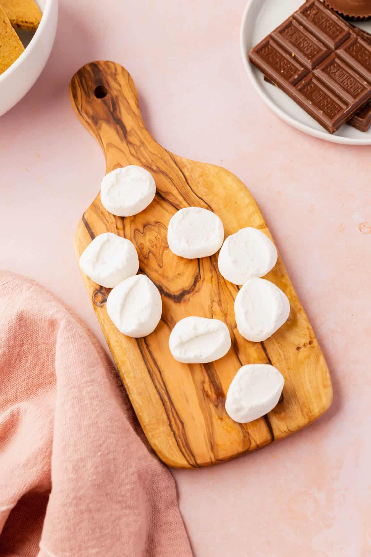 A small olivewood cutting board with eight marshmallows halves on it with a plate of chocolate bars on the side.