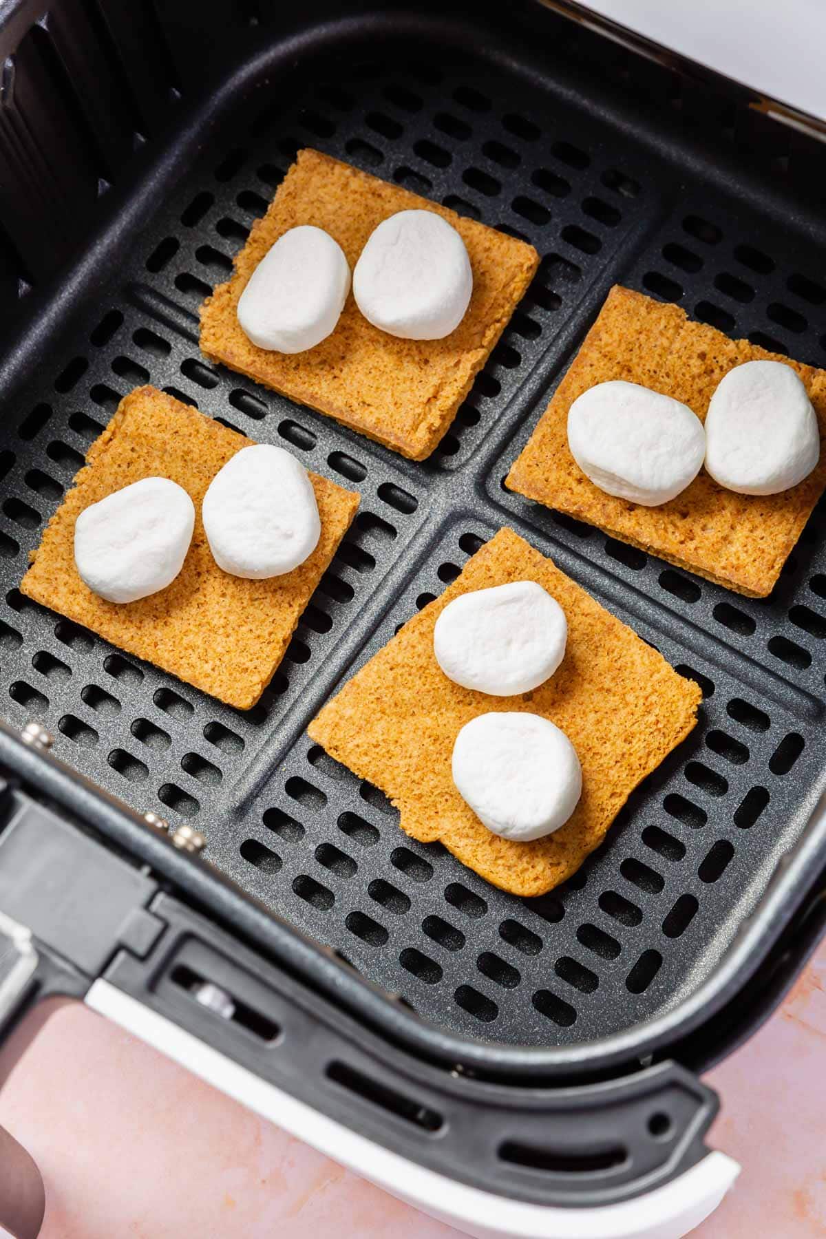 An air fryer basket with four graham cracker squares topped with two marshmallow halves each.