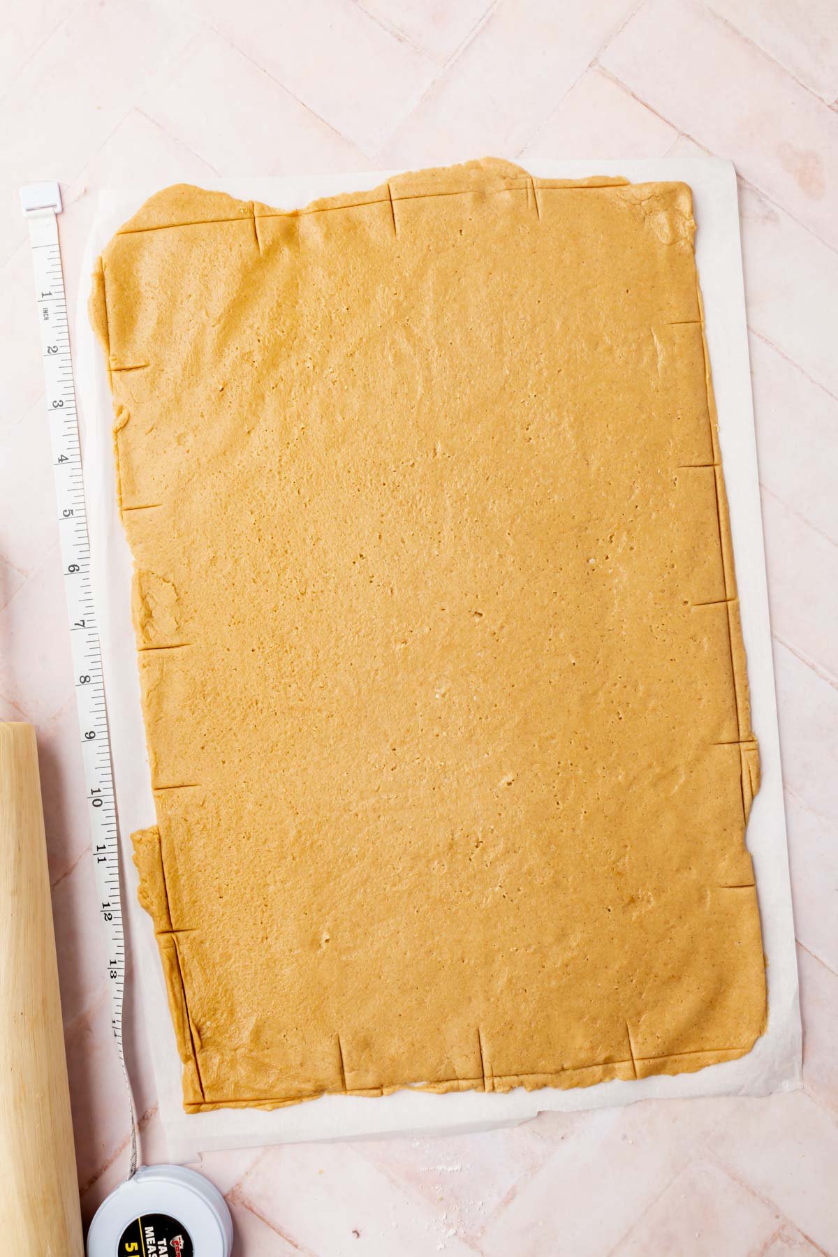 Graham cracker dough rolled out onto a piece of parchment paper with a measuring tape and a rolling pin next to it.