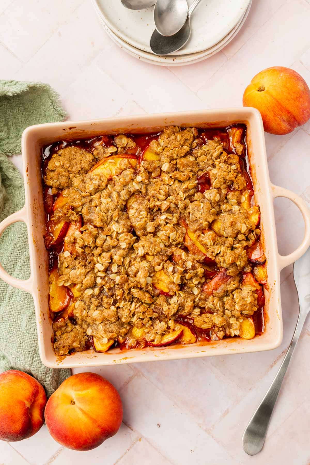 A square baking dish with gluten-free peach crisp surrounded by fresh peaches on a pink surface.