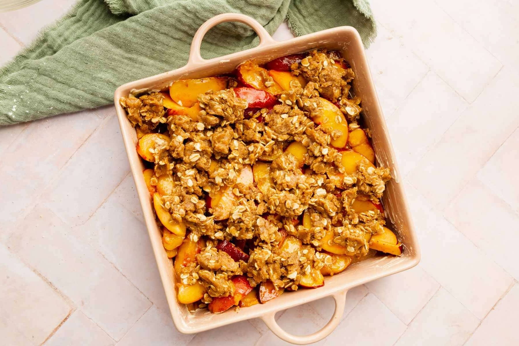 Unbaked peach crisp in a square baking dish with handles on a pink herringbone surface.