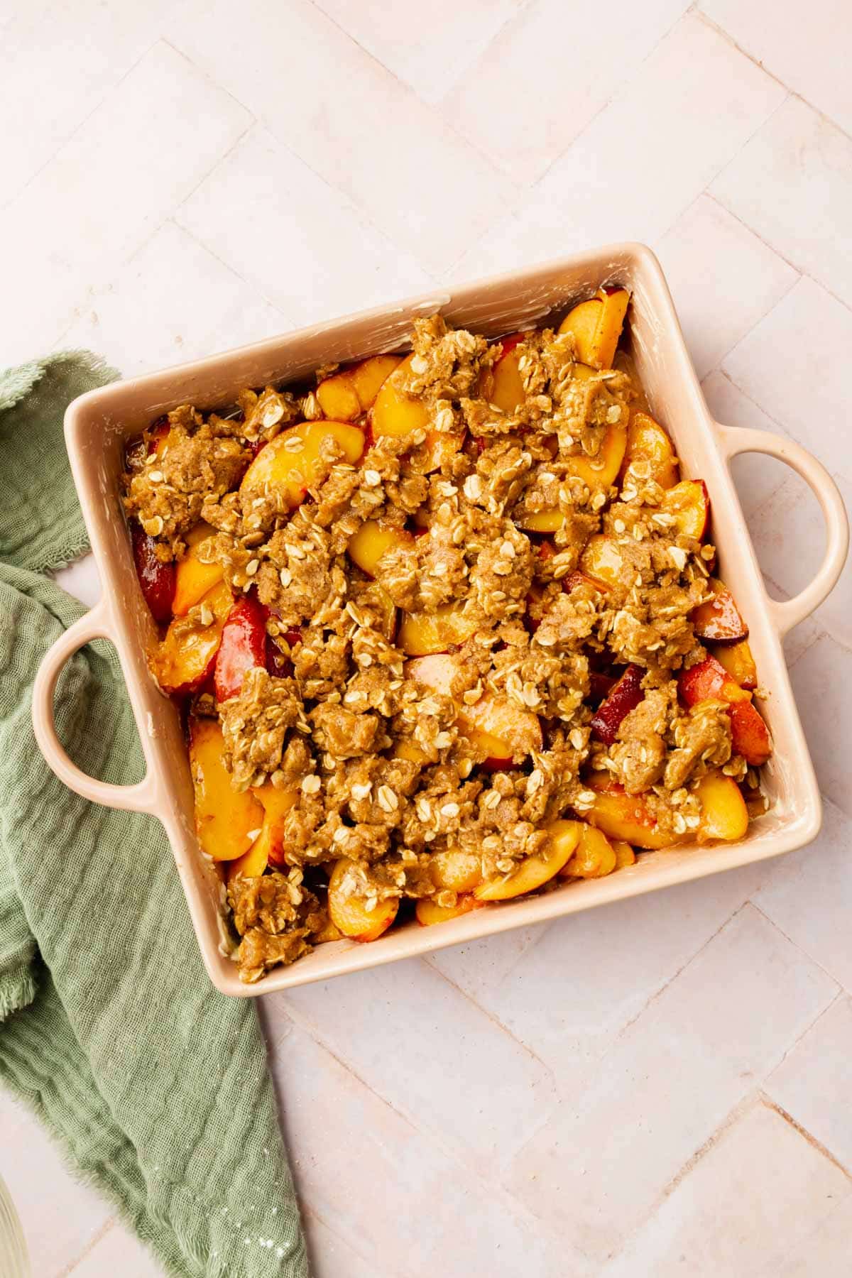 Unbaked peach crisp in a square baking dish with handles on a pink herringbone surface.