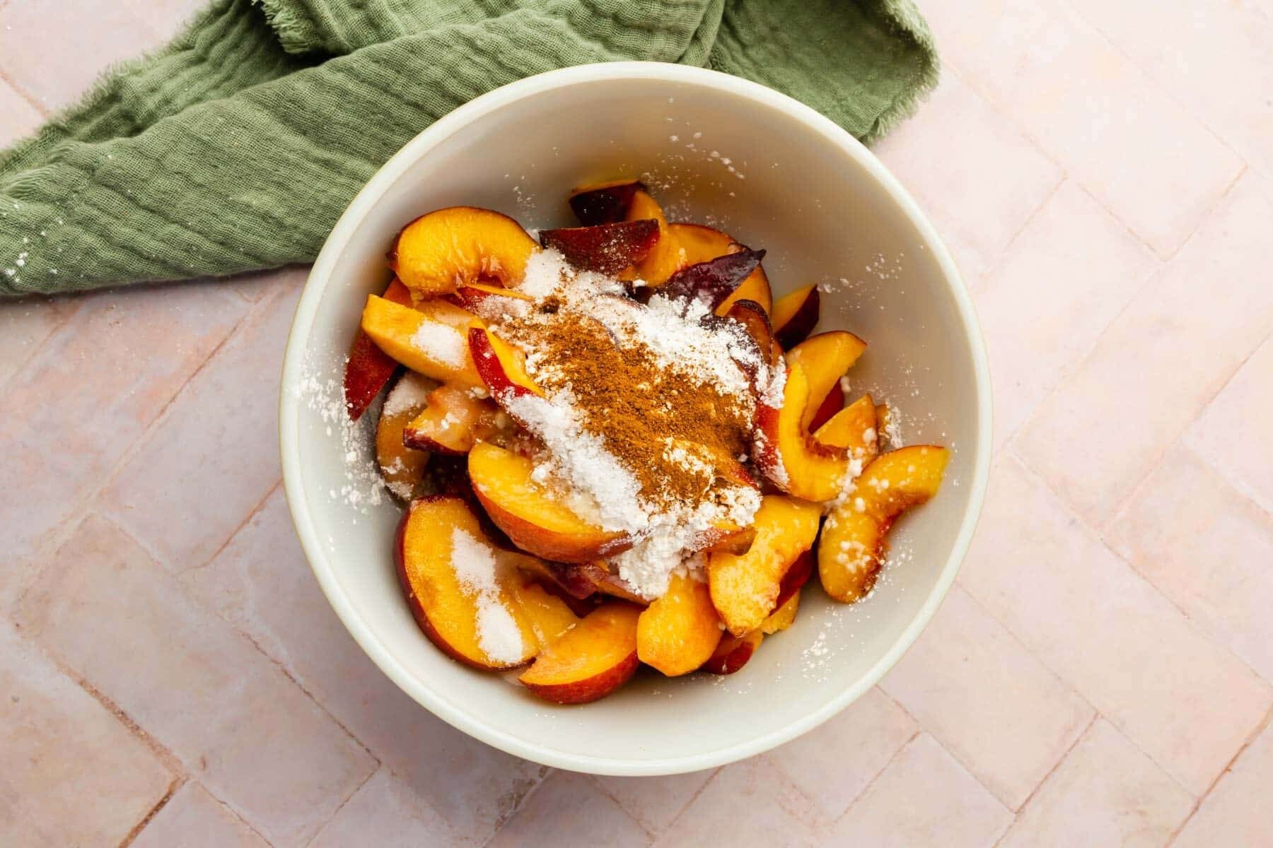 A bowl of sliced peaches, cornstarch, cinnamon, lemon juice, nutmeg, and granulated sugar ready to be mixed together.