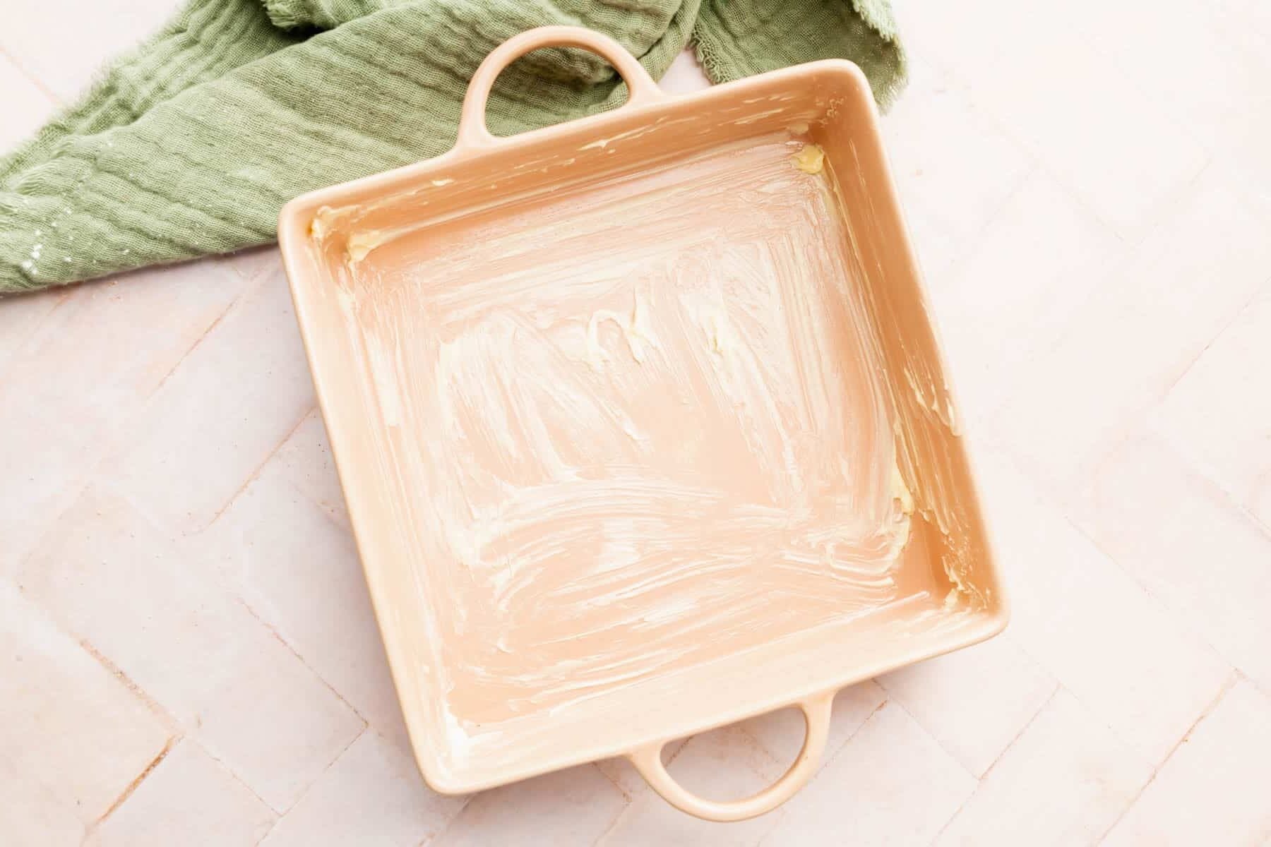 A buttered square baking dish with handles on top of a pink surface.