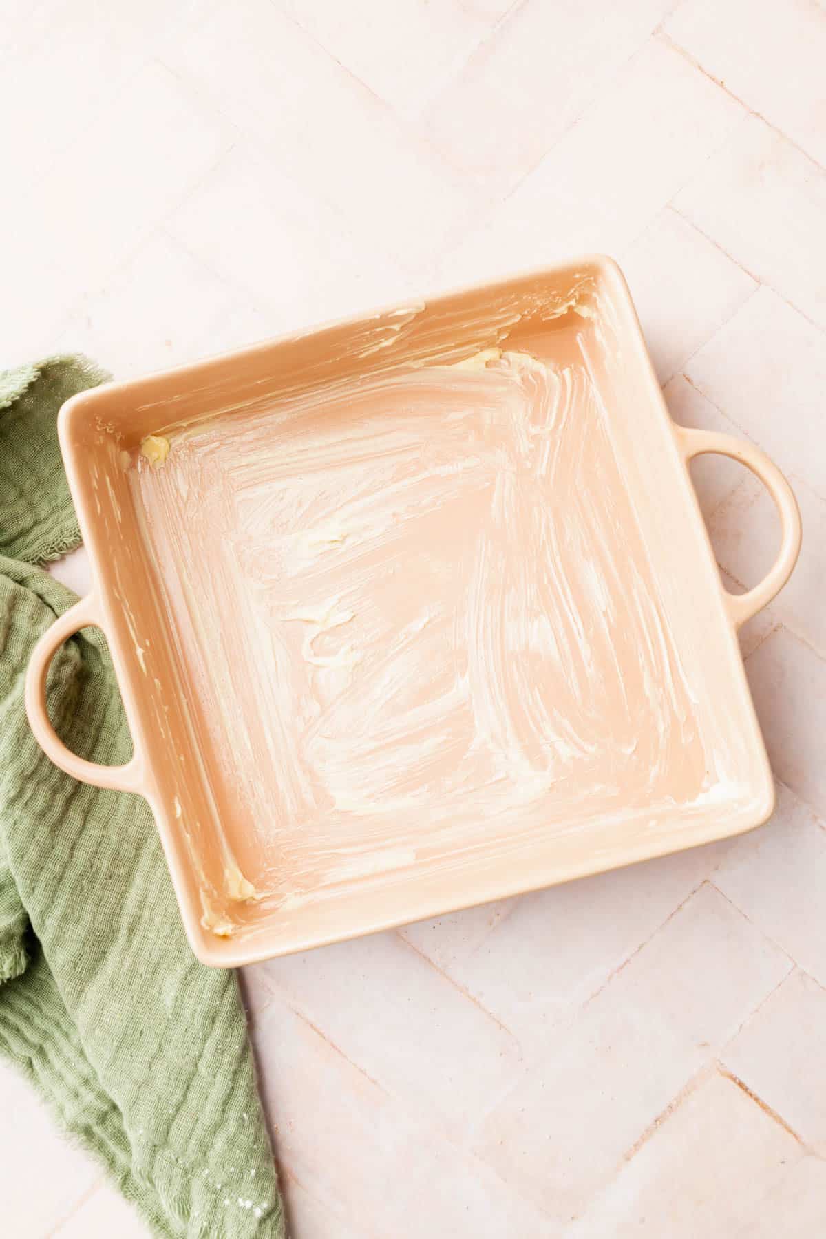 A buttered square baking dish with handles on top of a pink surface.