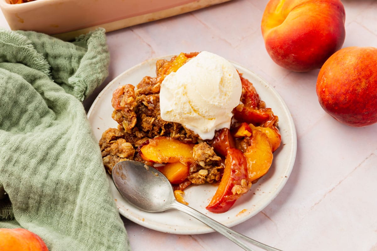 A single serving of peach crisp on a dessert plate with a single scoop of vanilla ice cream with a spoon and surrounded by fresh peaches.