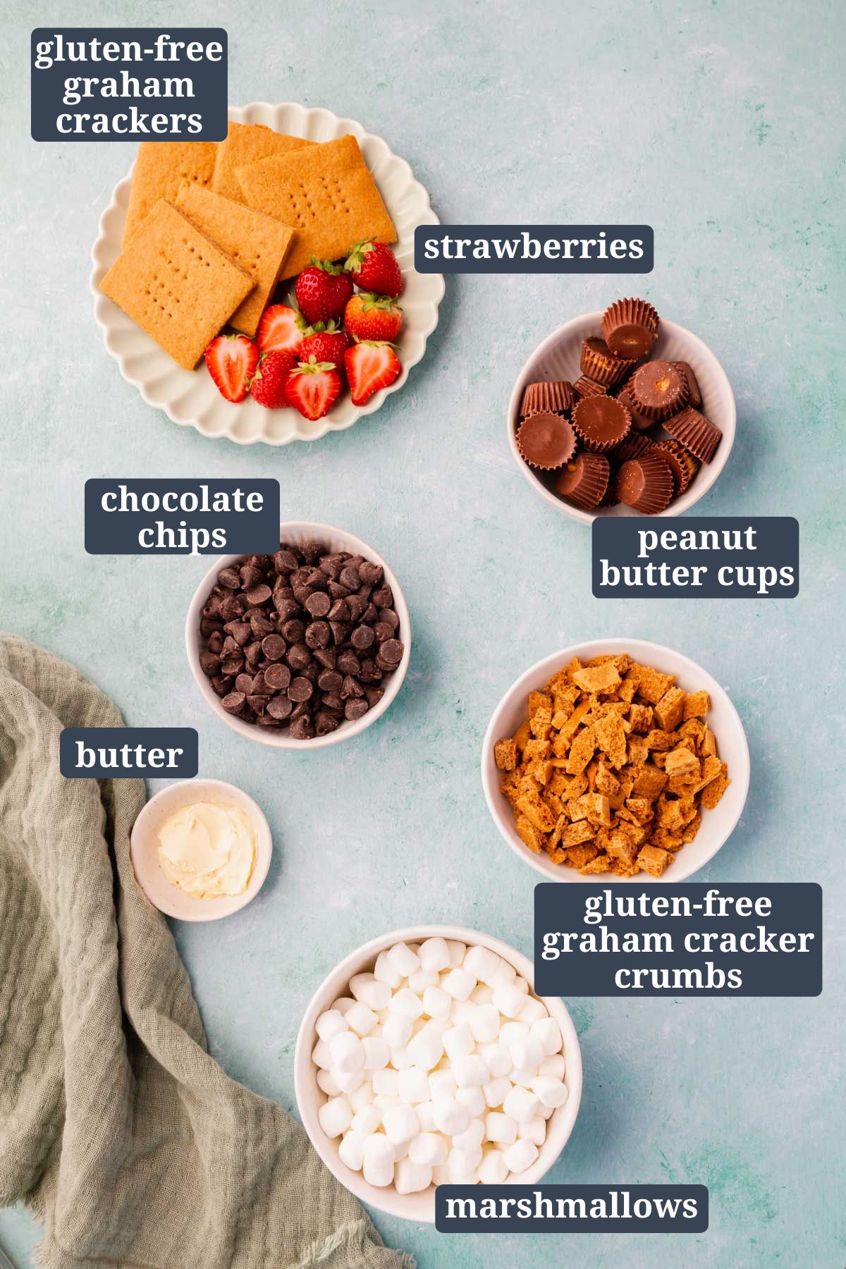 Small bowls of peanut butter cups, chocolate chips, gluten-free graham cracker crumbs, butter, mini marshmallows, graham crackers and strawberries for making s'mores dip on a blue background.