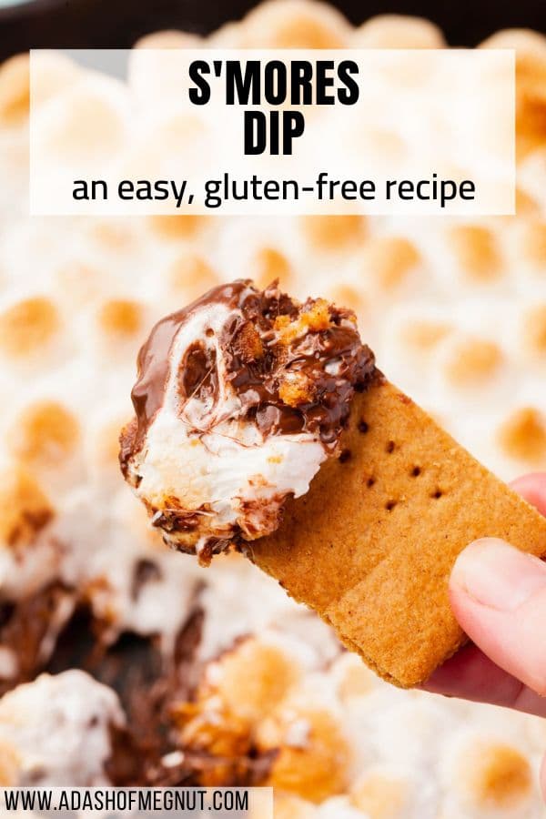A hand holding a gluten-free graham cracker dipped in s'mores dip held over the skillet of s'mores dip with a text overlay.