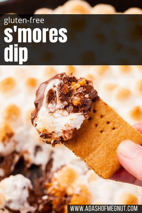 A hand holding a graham cracker topped with s'mores dip over a skillet of s'mores dip.