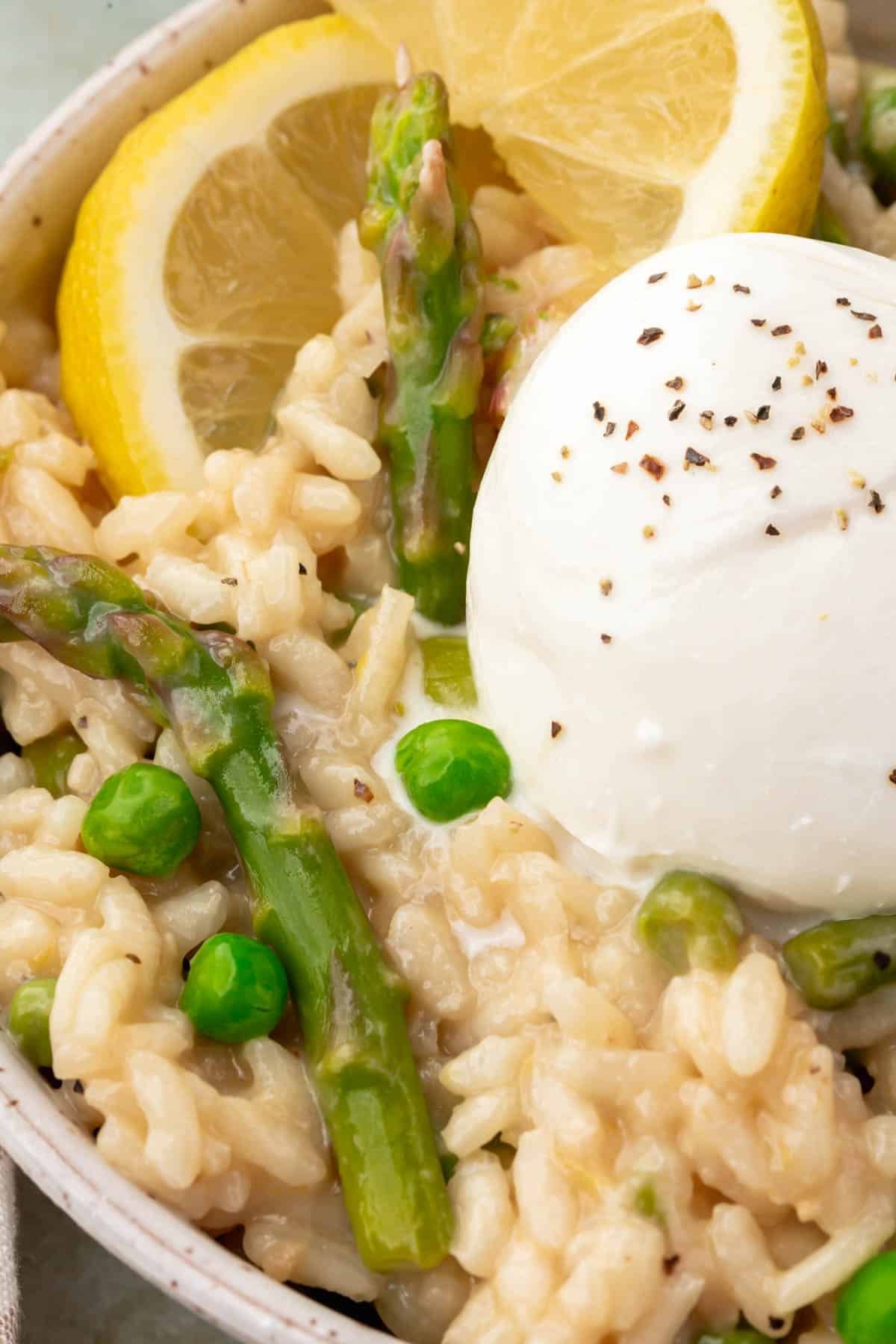 A closeup of a bowl of risotto with peas and asparagus topped with a ball of burrata sprinkled with black pepper and a lemon wedge.