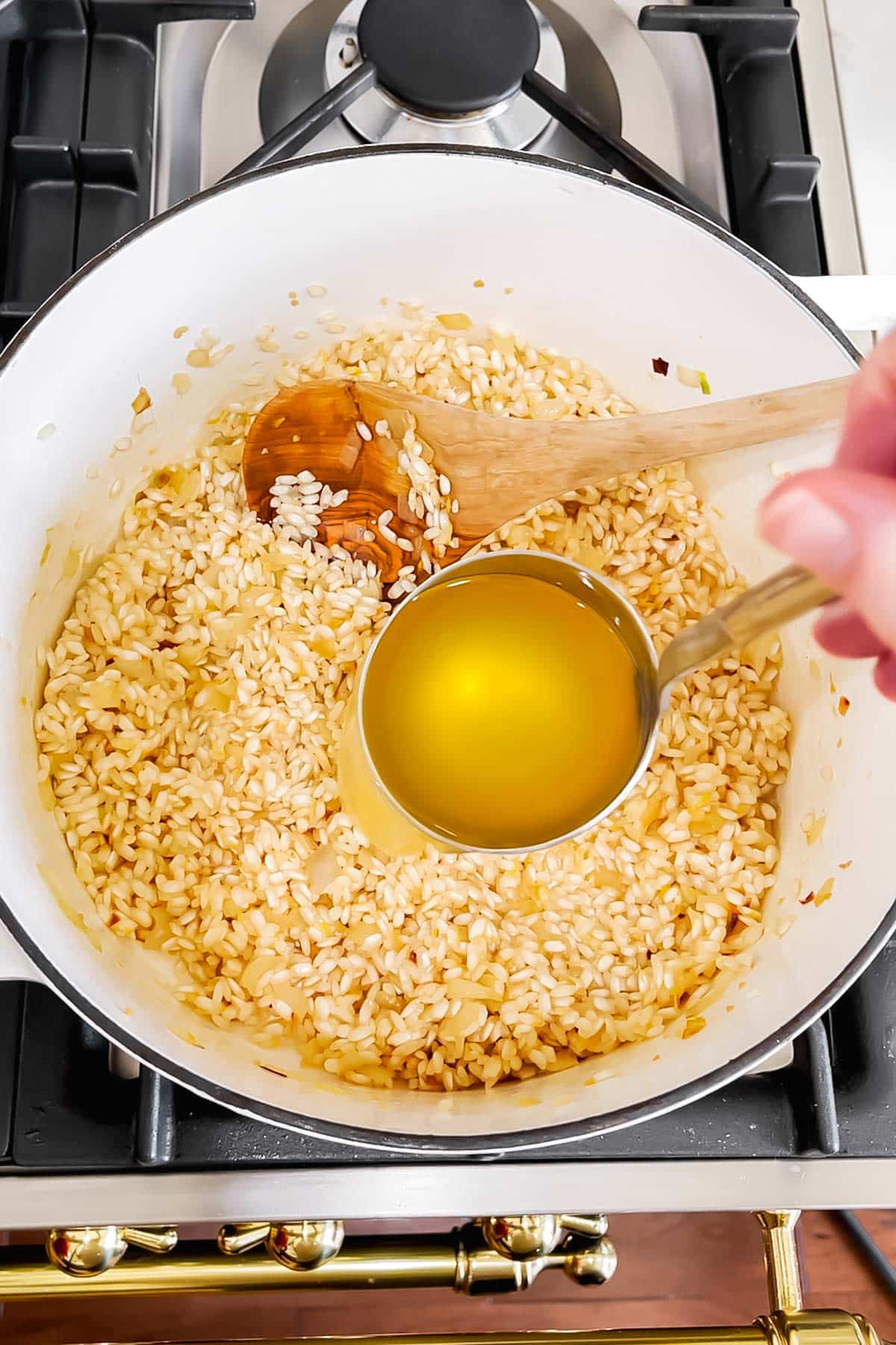 A ladle of vegetable broth is being added to arborio rice in a dutch oven to make risotto.