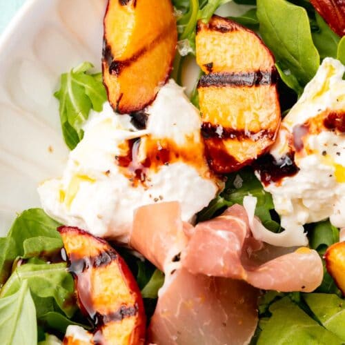 A closeup of a burrata grilled peach salad topped with arugula, basil and prosciutto on a white platter.