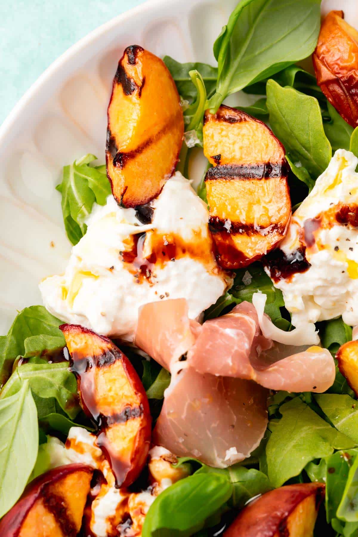 A closeup of a burrata grilled peach salad topped with arugula, basil and prosciutto on a white platter.