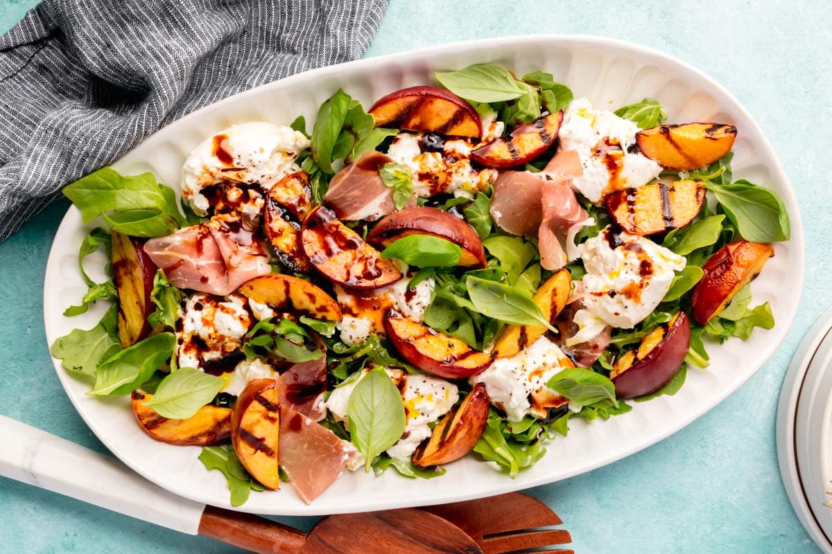 An oval plate with arugula grilled peach burrata salad topped with prosciutto and balsamic glaze on a blue table with wood salad tongs.