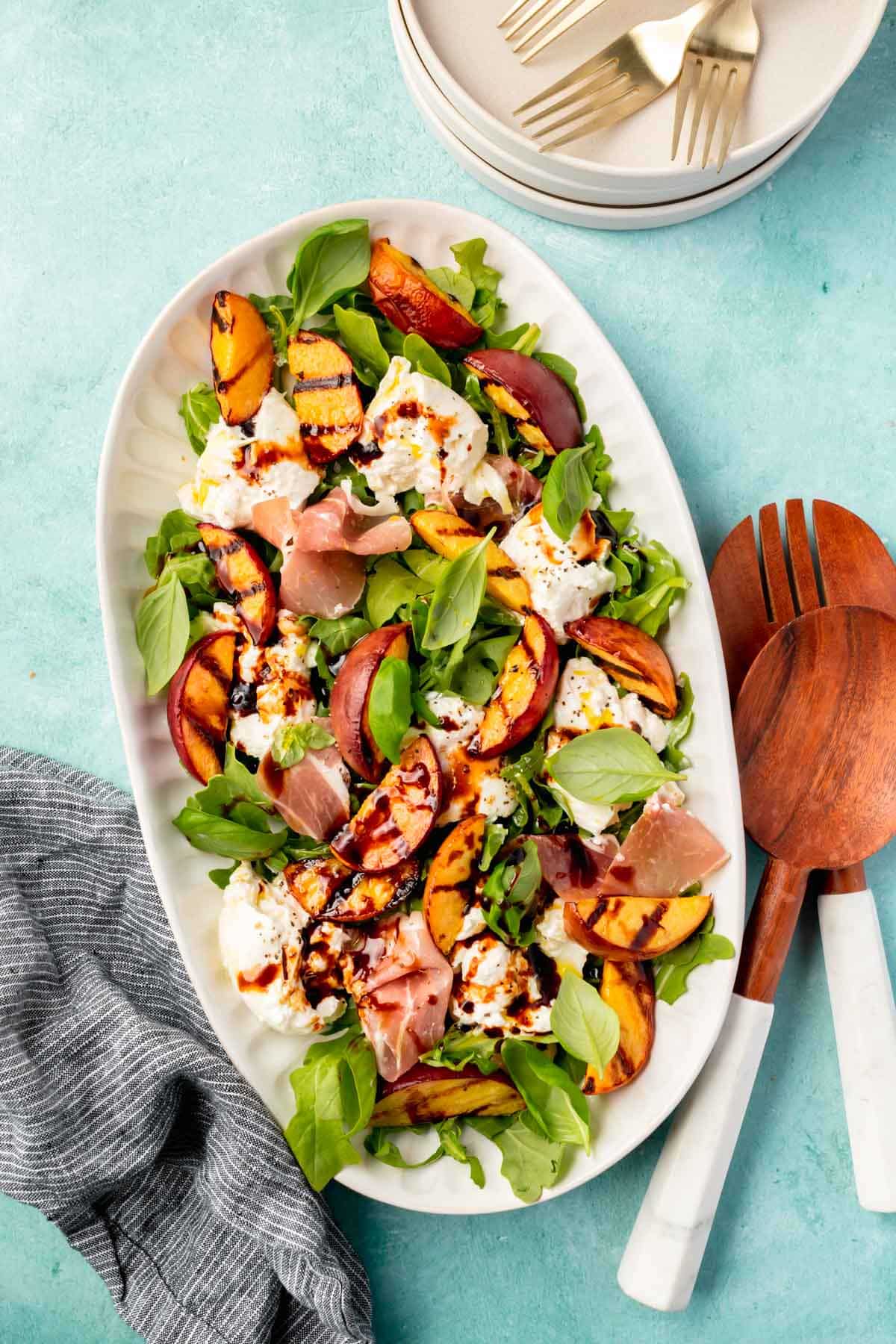 An oval platter topped with grilled peach burrata salad topped with basil, prosciutto, arugula, balsamic glaze and black pepper on a blue table with a dark blue napkin.