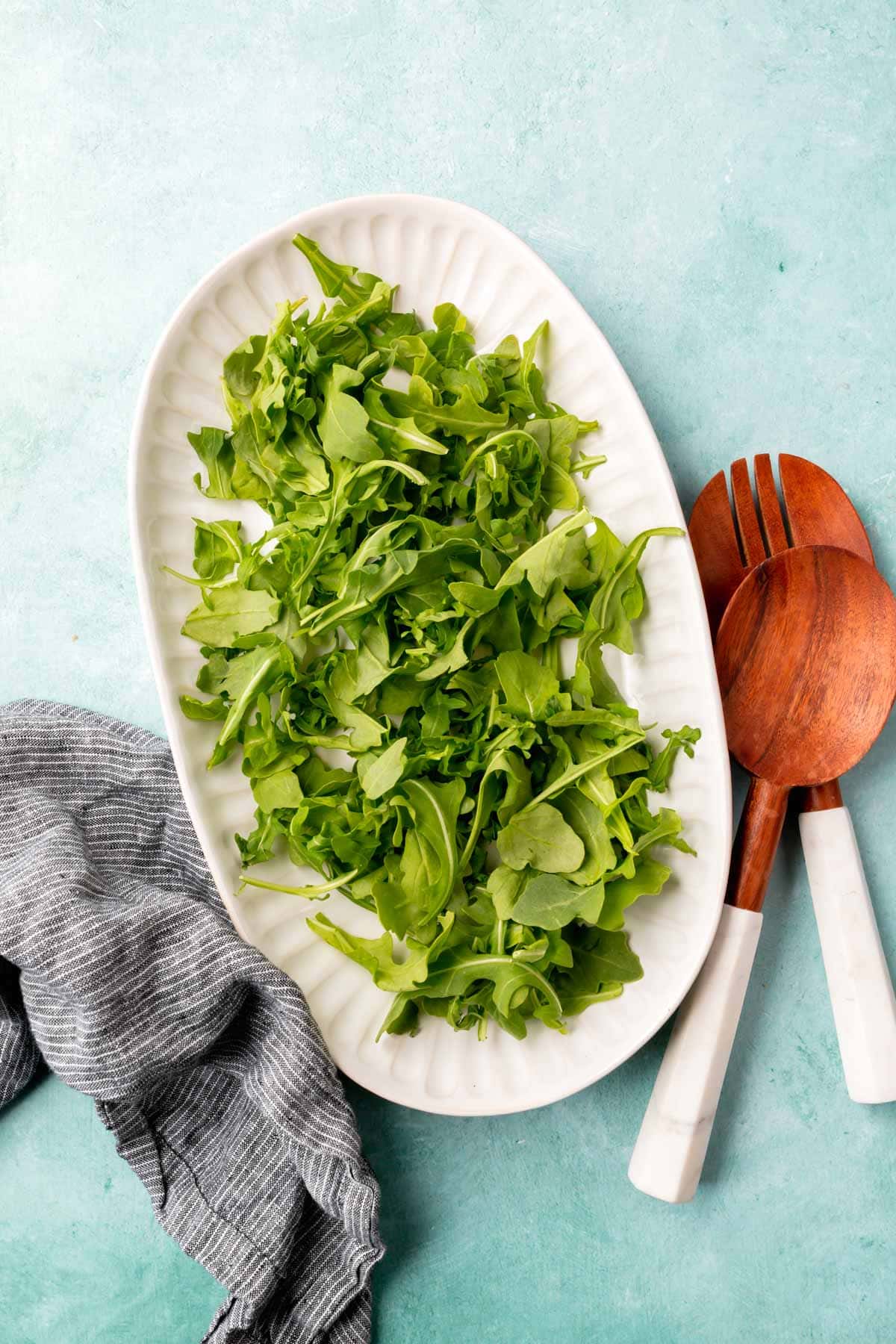 A large oval platter of arugula sitting on a bright blue table with wood and marble salad tongs to the side.
