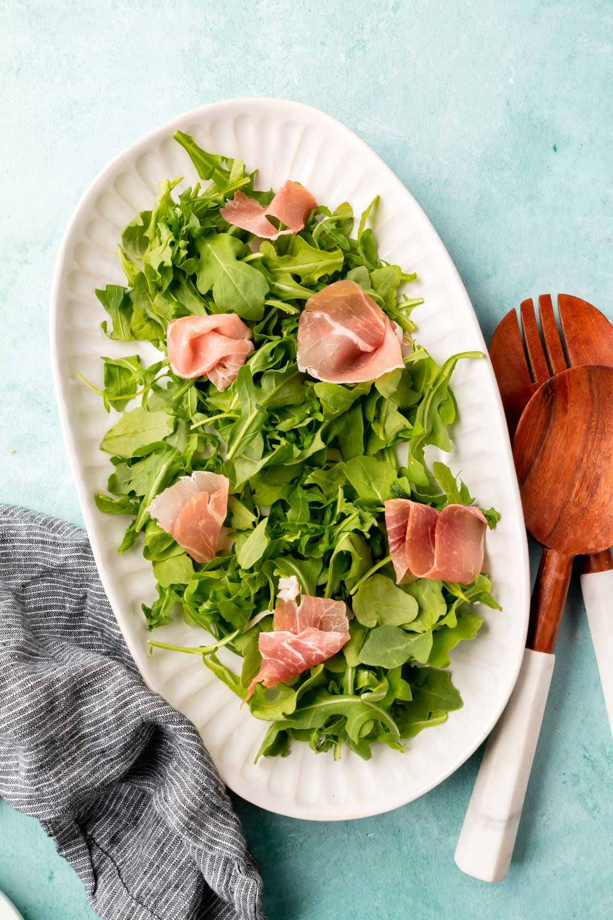 A large oval platter on a blue table topped with arugula and prosciutto with wood and marble salad tongs to the side.