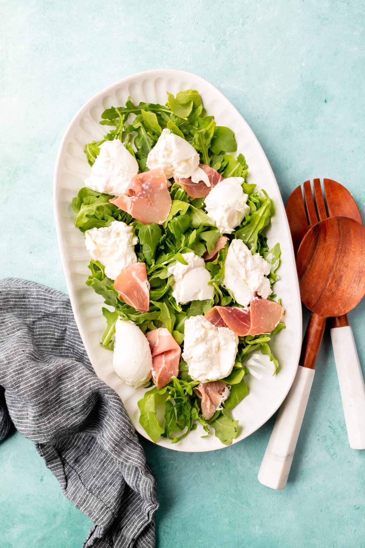 A large oval platter on a bright blue table topped with arugula, burrata and prosciutto with salad tongs on the side.