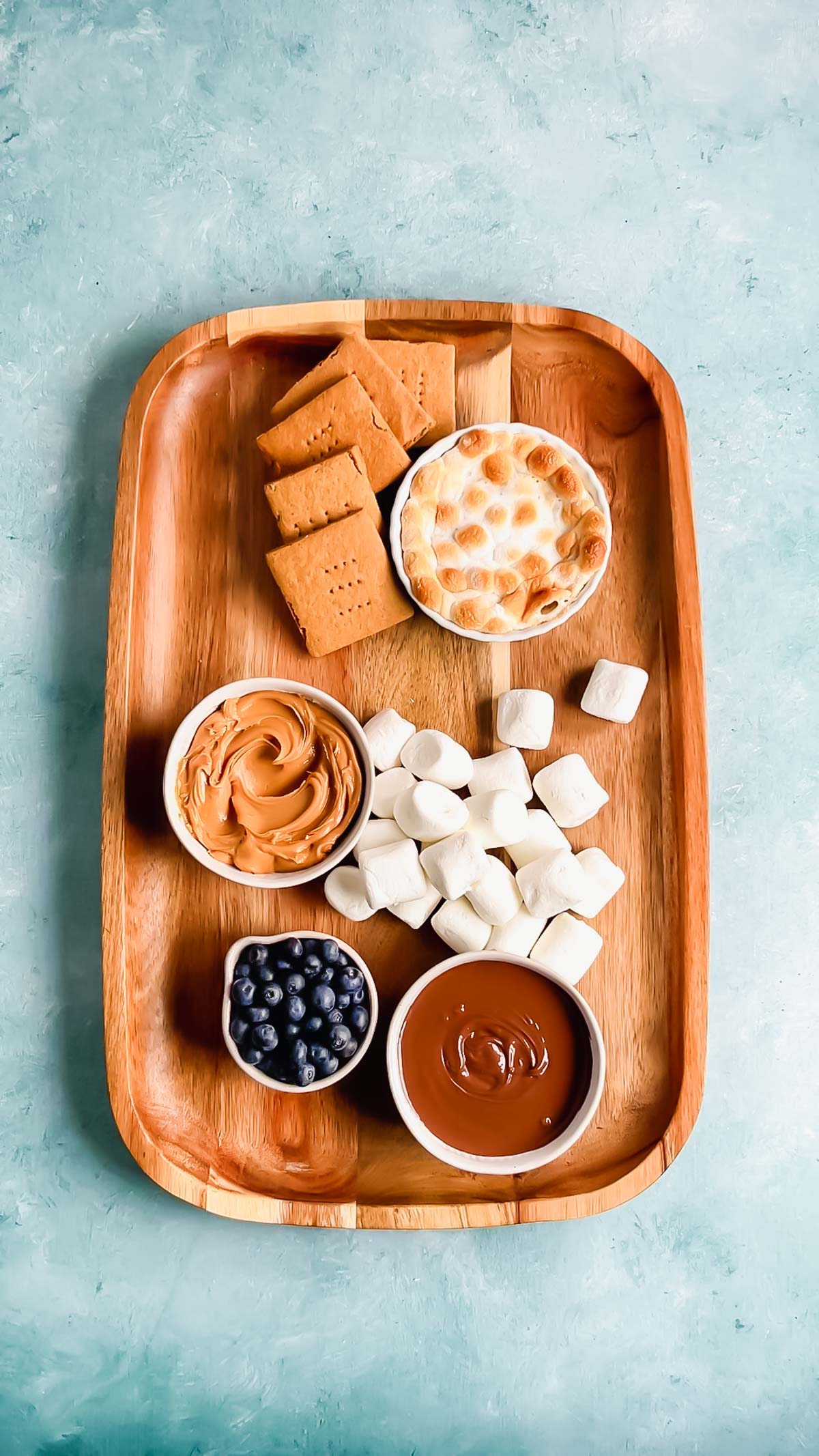 A large wooden rectangular chacuterie board with marshmallows, graham crackers, and bowls of nutella, s'mores dip, peanut butter, and blueberries.