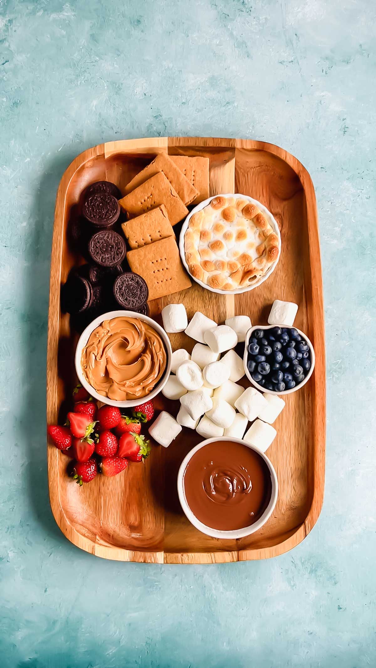 A large wooden rectangular charcuterie board with glute-free Oreos, graham crackers, marshmallows, strawberries, and bowls of s'mores dip, peanut butter, blueberries and nutella.