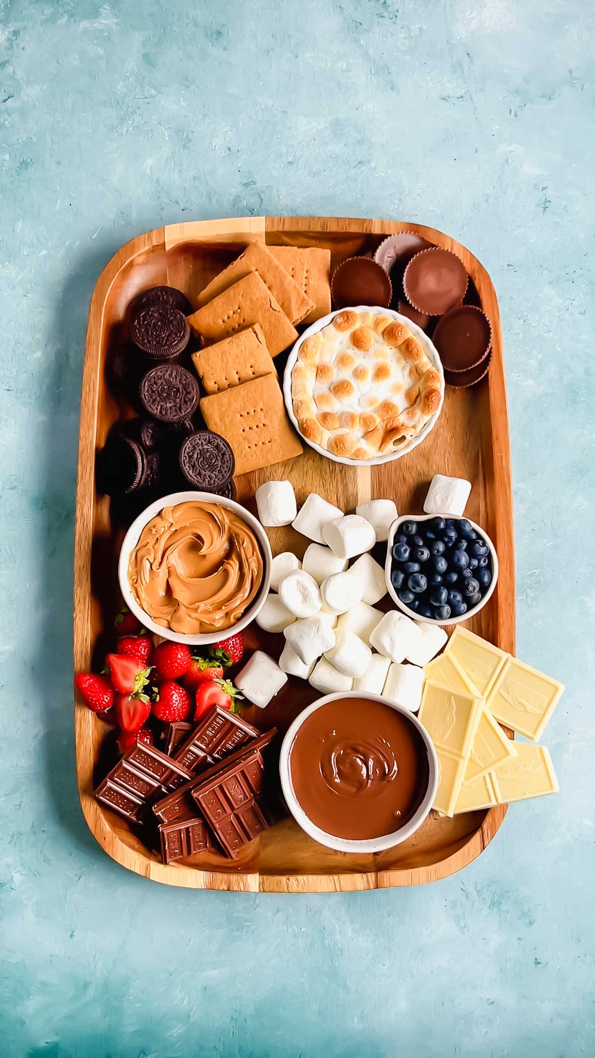 A large wooden rectangular board with gluten-free Oreos, graham crackers, peanut butter cups, marshmallows, strawberries, white chocolate bars, milk chocolate bars, and bowls of nutella, peanut butter, blueberries, and s'mores dip.