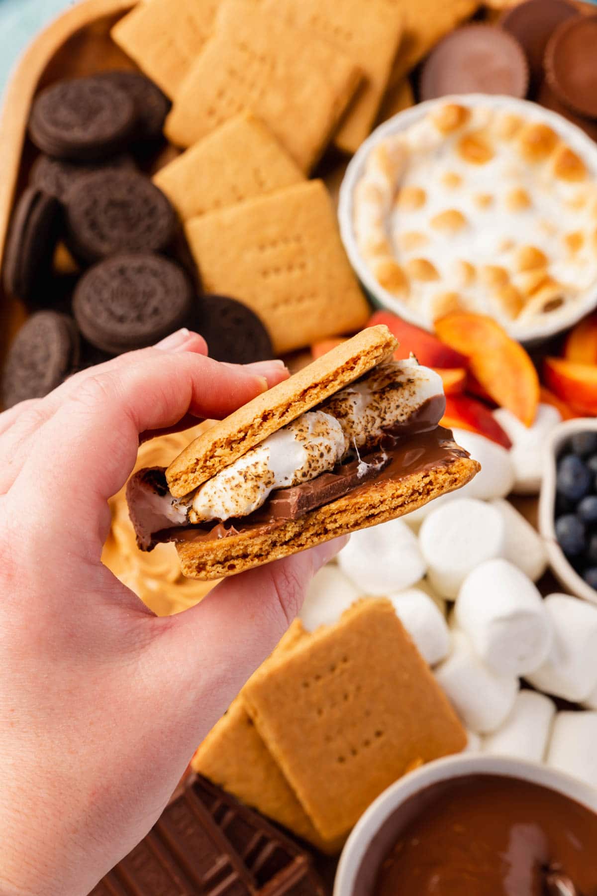 A hand holding a gluten-free s'mores over a s'mores charcuterie board.