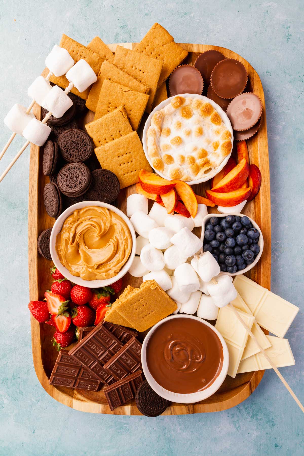 A s'mores charcuterie board with marshmallows, oreos, gluten-free graham crackers, s'mores dip, nutella, peanut butter, strawberries, blueberries and peaches on a blue background.