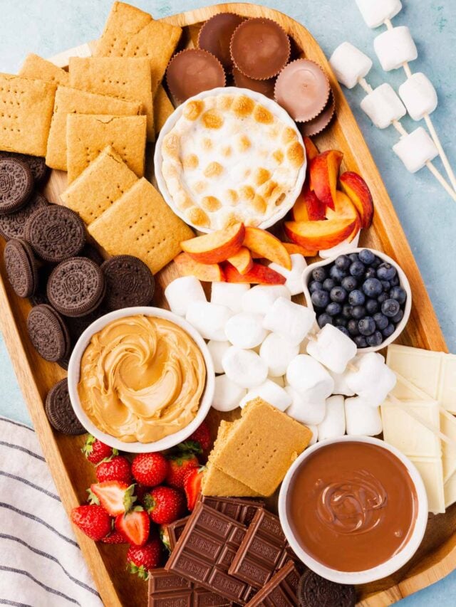 A s'mores charcuterie board filled with marshmallows, chocolate, graham crackers, fruit, and dips on a blue surface.