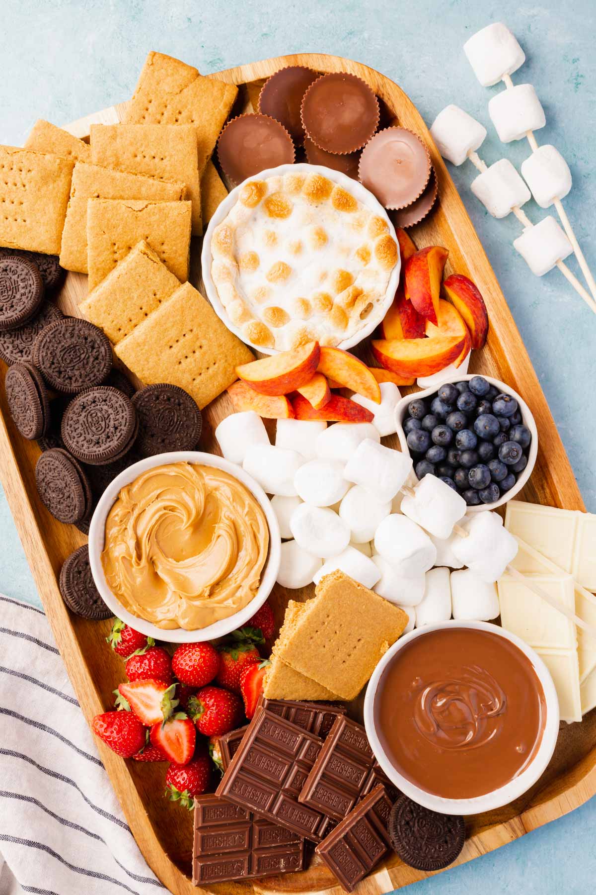 A s'mores charcuterie board filled with marshmallows, chocolate, graham crackers, fruit, and dips on a blue surface. 