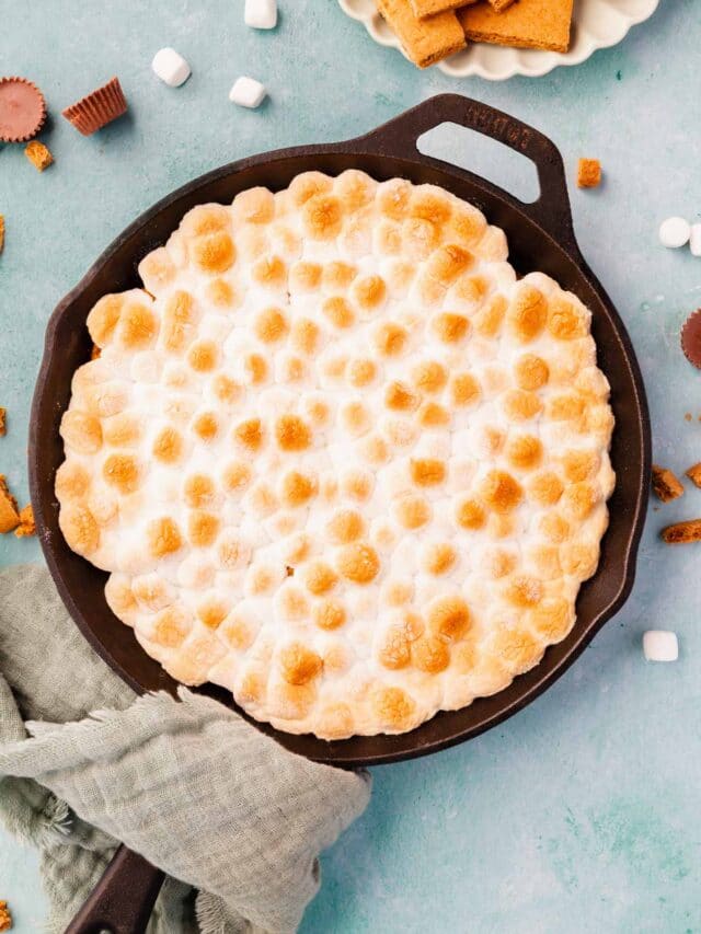 A cast iron skillet filled with toasted marshmallow s'mores dip on a blue background with a green napkin wrapepd around the handle.