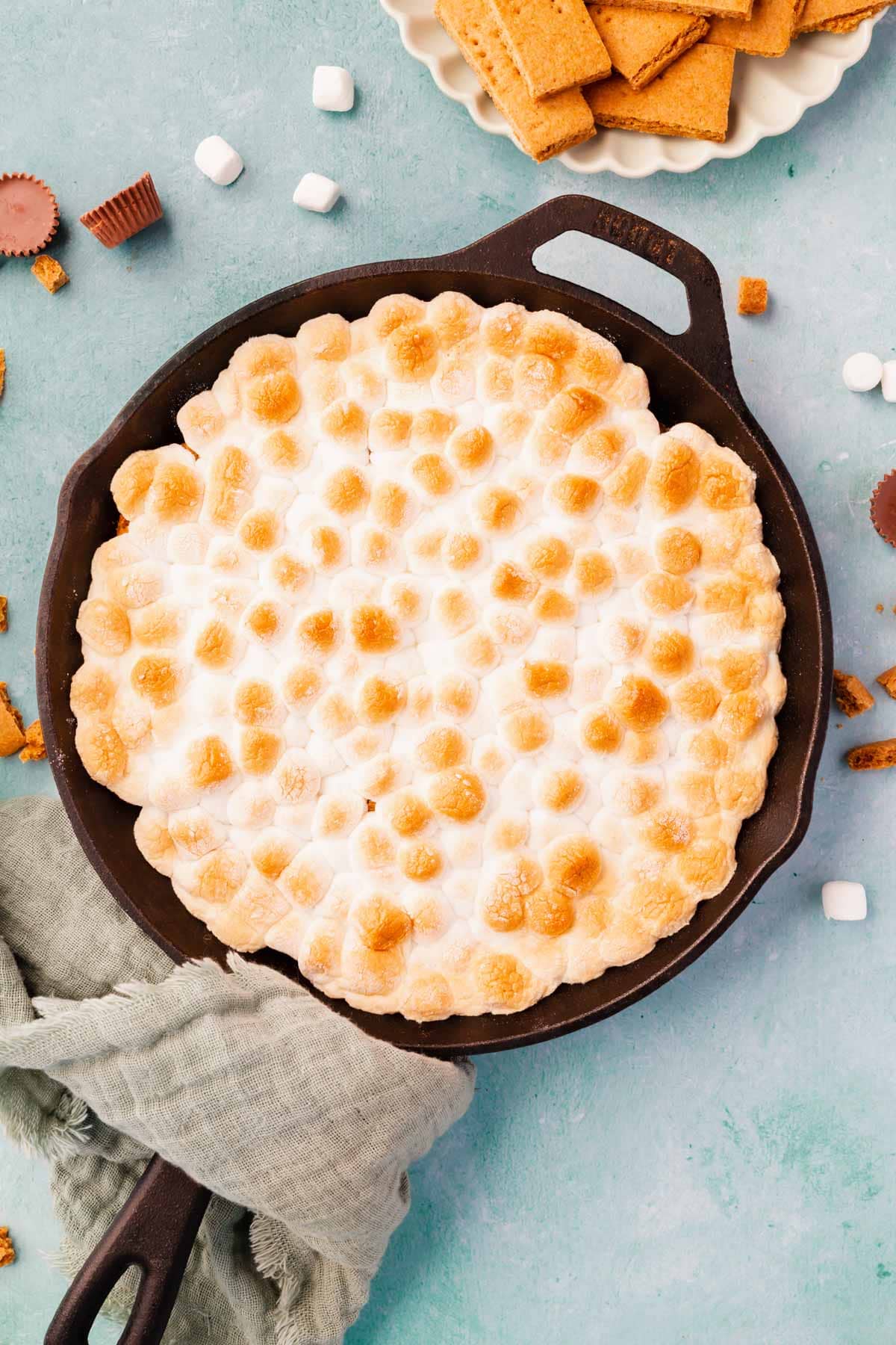 A cast iron skillet filled with toasted marshmallow s'mores dip on a blue background with a green napkin wrapepd around the handle.