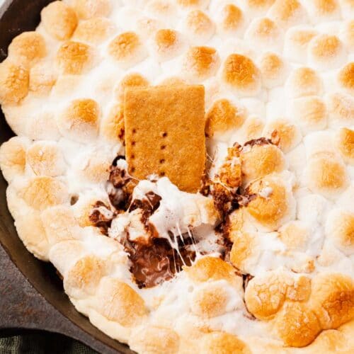 A gluten-free graham cracker being dipped into s'mores dip in a cast iron skillet.