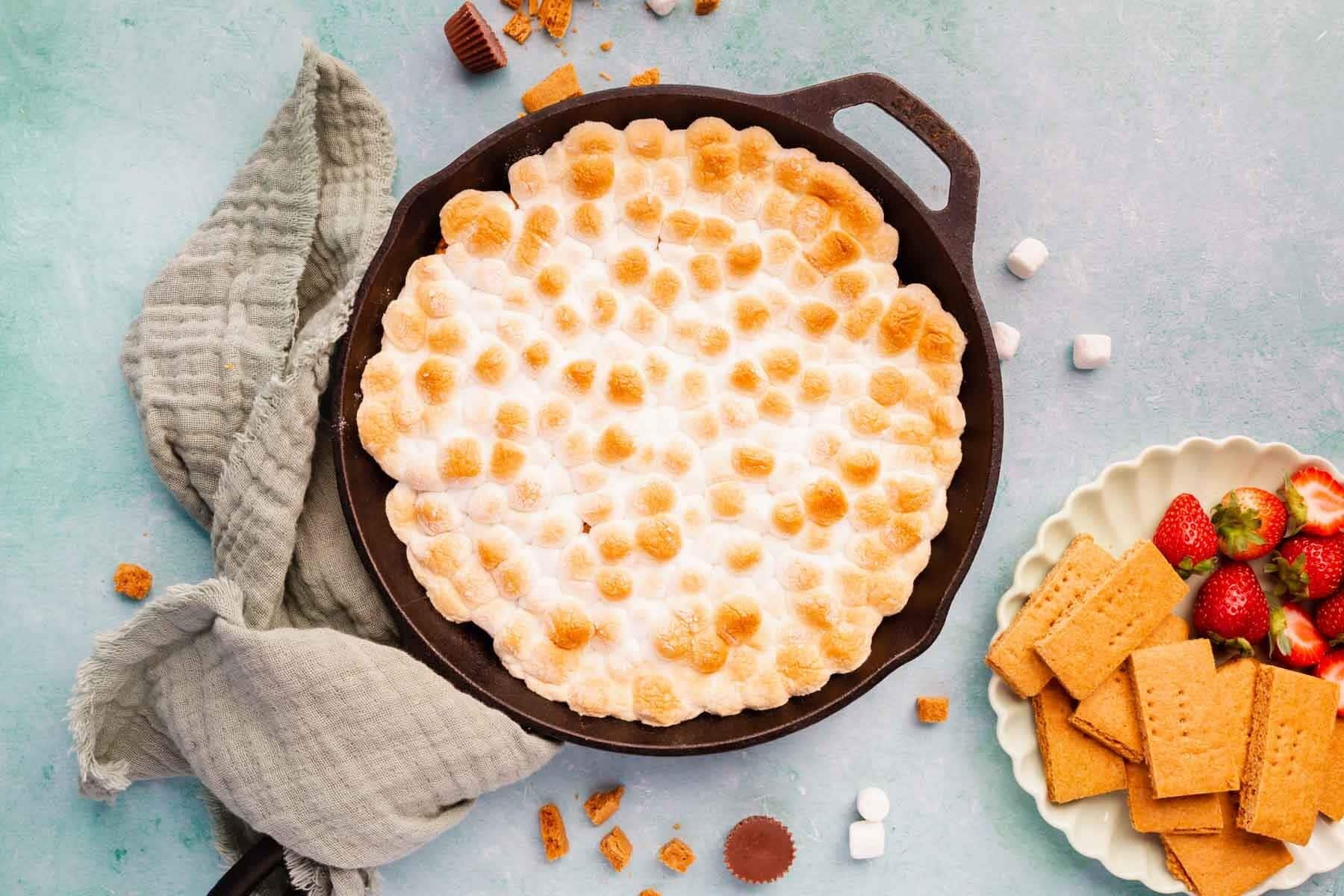 Toasted marshmallows over s'mores dip in a cast iron skillet with a green linen napkin on a blue background with a plate of graham crackers and strawberries.