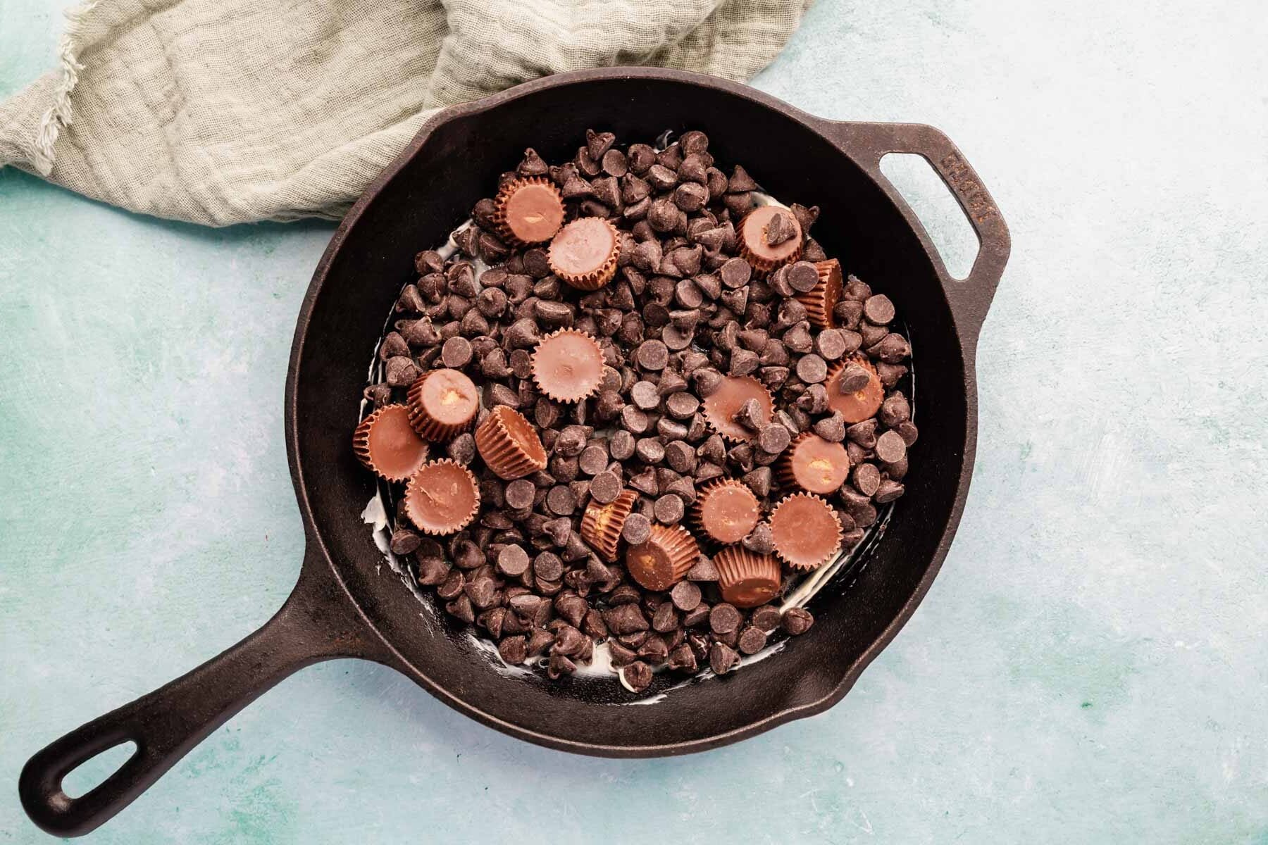 Chocolate chips and peanut butter cups layered in a cast iron skillet on a blue background.