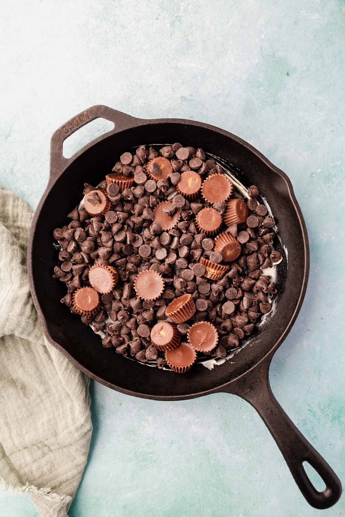 Chocolate chips and peanut butter cups layered in a cast iron skillet on a blue background.