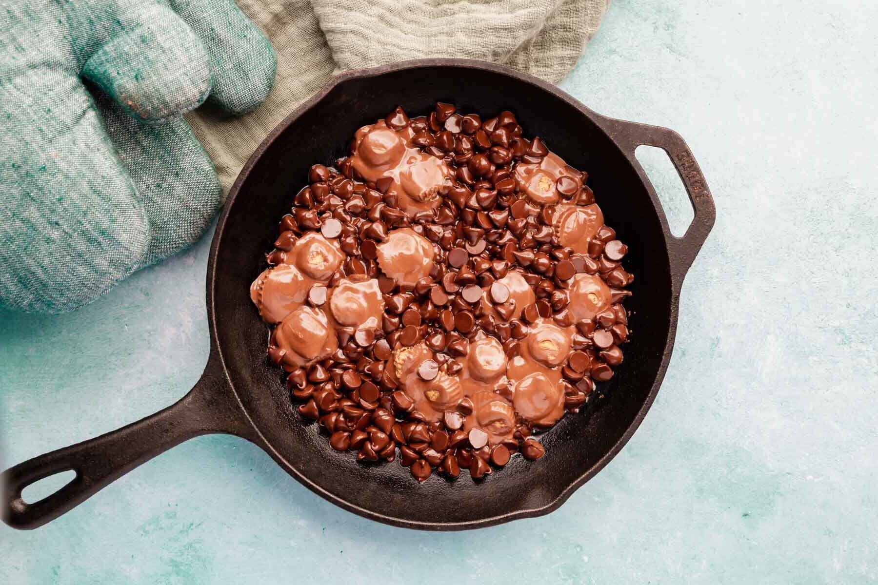 Glossy chocolate chips and peanut butter cups starting to melt in a cast iron skillet on a blue background.