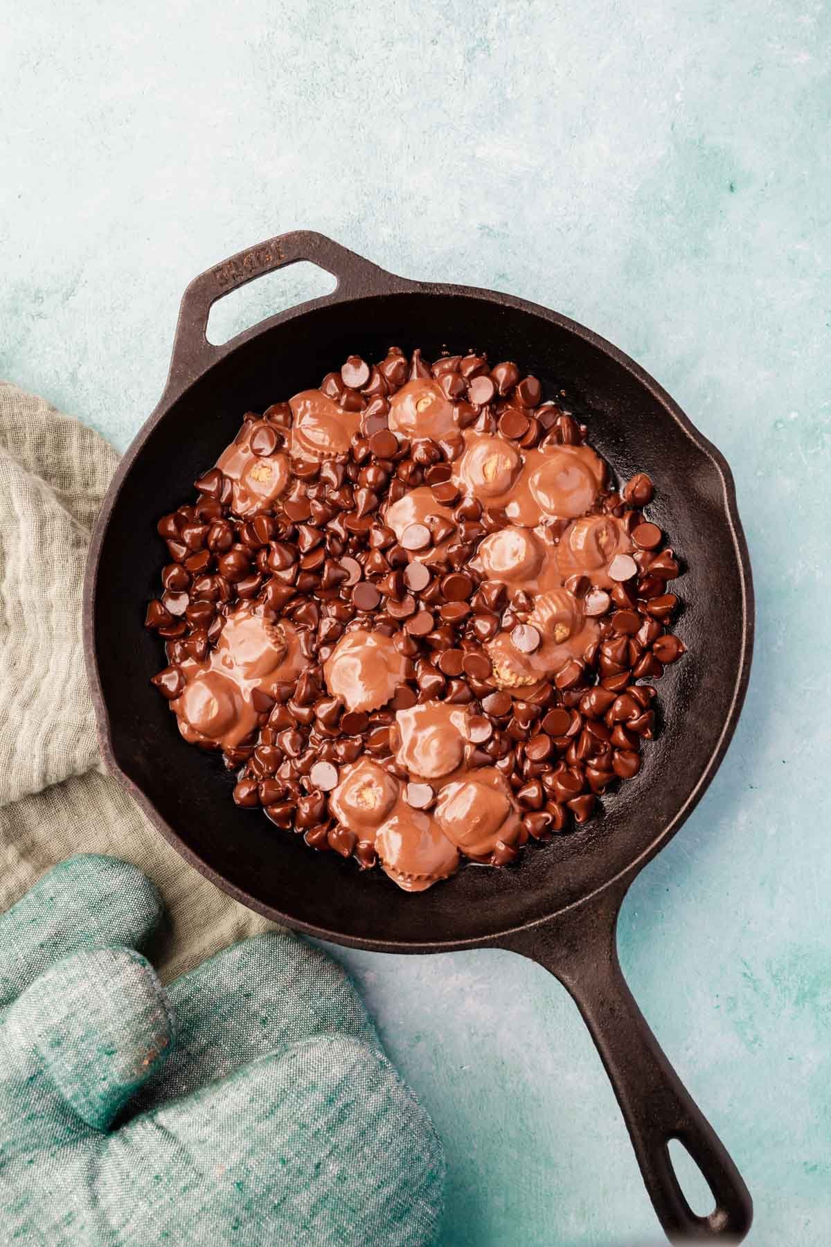Glossy chocolate chips and peanut butter cups starting to melt in a cast iron skillet on a blue background.