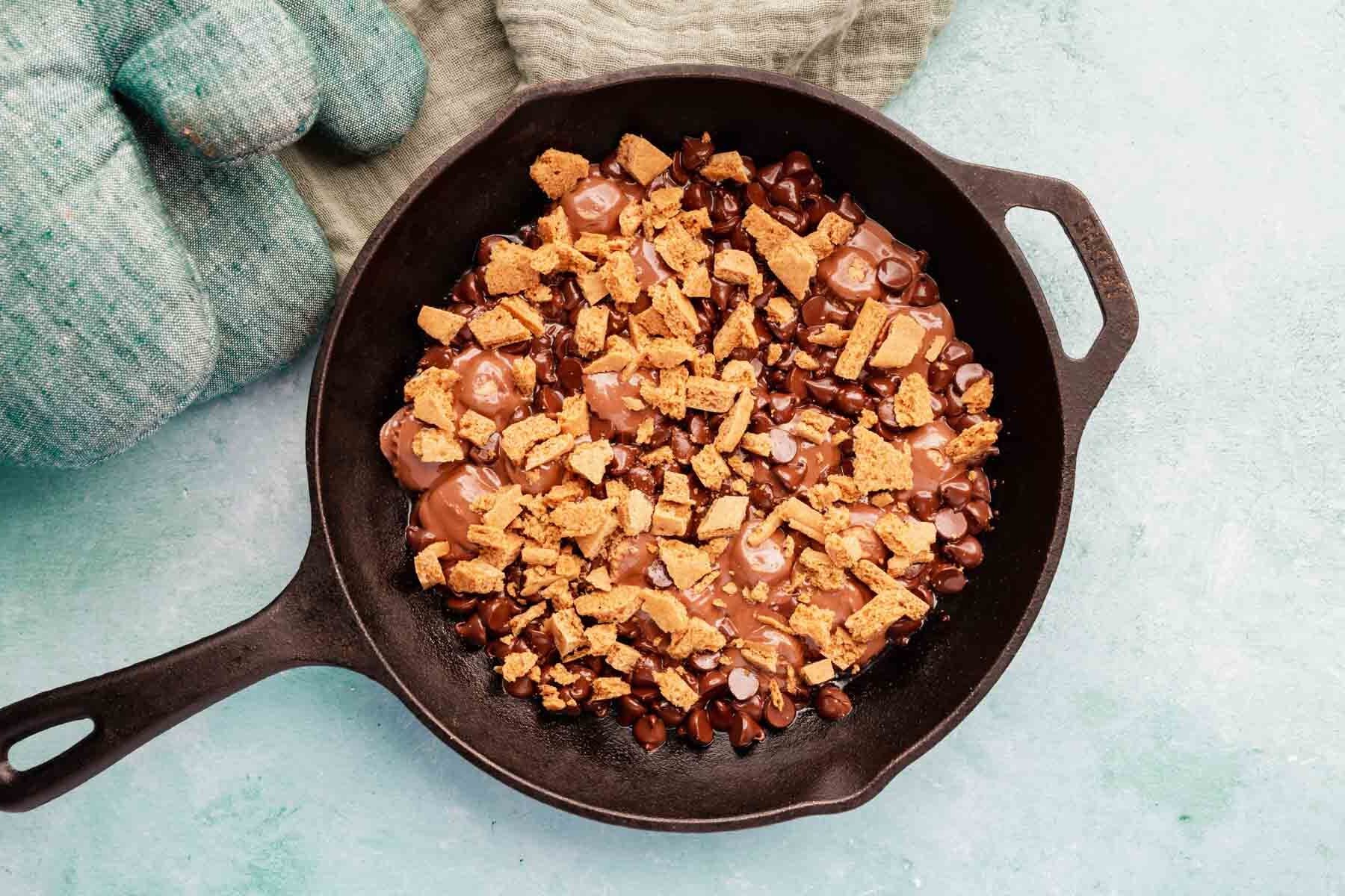 Graham cracker crumbs layered over melted chocolate chips and peanut butter cups in a cast iron skillet.