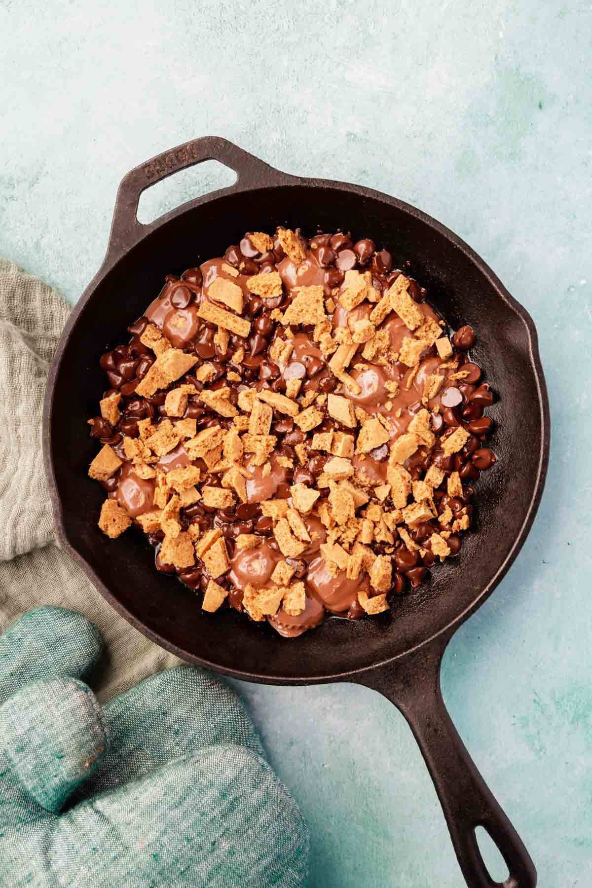 Graham cracker crumbs layered over melted chocolate chips and peanut butter cups in a cast iron skillet.